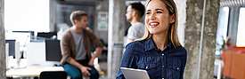 Eine lächelnde Frau hält ein Tablet in der Hand und steht in einem modernen Büro. Im Hintergrund sind zwei Männer zu sehen, die miteinander sprechen. Die Atmosphäre wirkt freundlich und kooperativ.