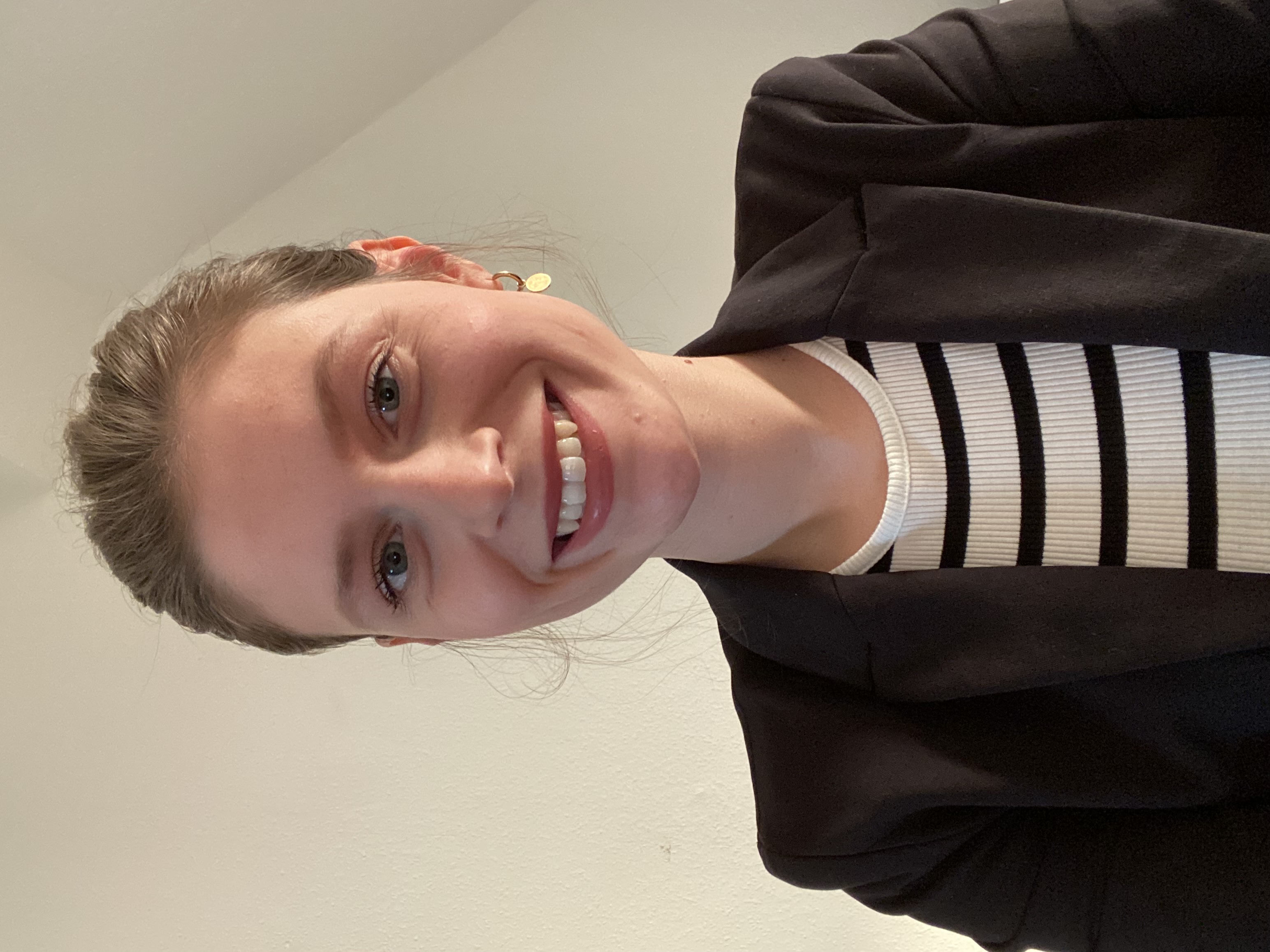 Eine lächelnde Frau mit langen, blonden Haaren trägt einen schwarzen Blazer und ein gestreiftes Oberteil. Sie sitzt vor einer hellen Wand und schaut in die Kamera.